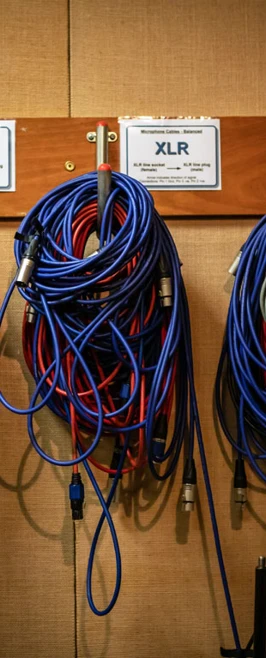 Various blue and black XLR cables neatly coiled and hung on a wooden rack, labeled for different microphone and speaker uses. Various blue and black XLR cables neatly coiled and hung on a wooden rack, labeled for different microphone and speaker uses.